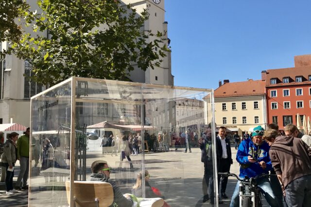 Zwei Personen sitzen im Glaskasten in der Regensburger Innenstadt