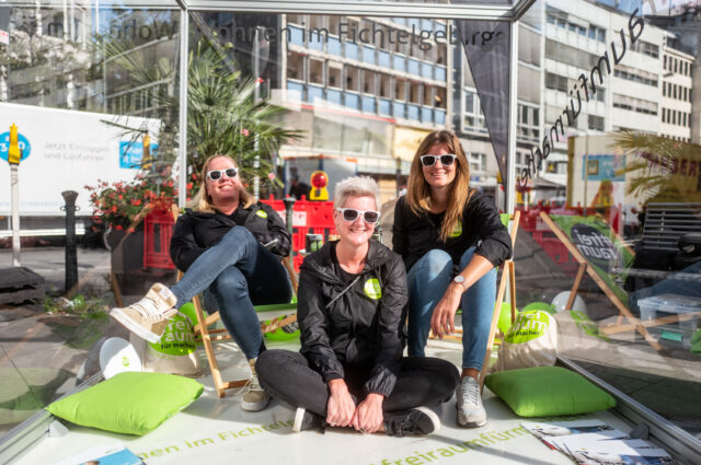 Drei Frauen sitzen im Aktionsglaskasten in der Düsseldorfer Innenstadt und lächeln fröhlich