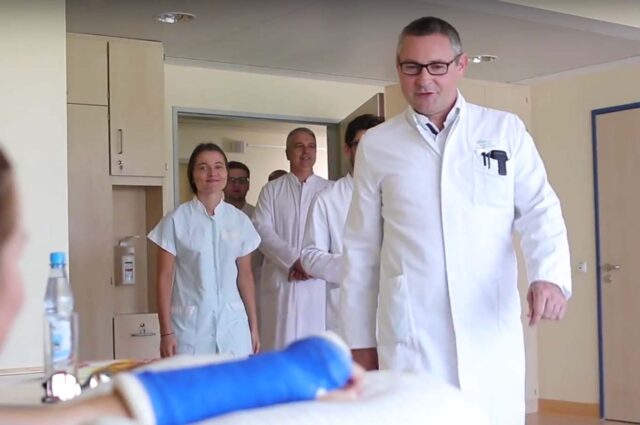 Arzt während einer Visiten bei einem Patienten mit mehreren Ärzten im Hintergrund die durch die Tür reinkommen