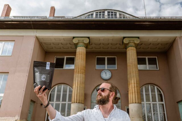 Mann mit Sonnenbrille und langen Haaren steht mit einer Packung Kaffee vor einem Gebäude