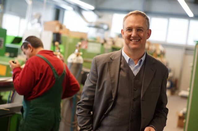 Jörg de Signier steht in einer Produktionshalle und lächelt mit einer Hand in der Hosentasche in die Kamera im Hintergrund arbeitet ein Mann in Latzhose