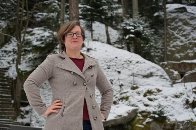 Birgit Simmler steht mit beiden Händen in den Hüften vor einer schneebedeckten hügeligen Landschaft und lächelt