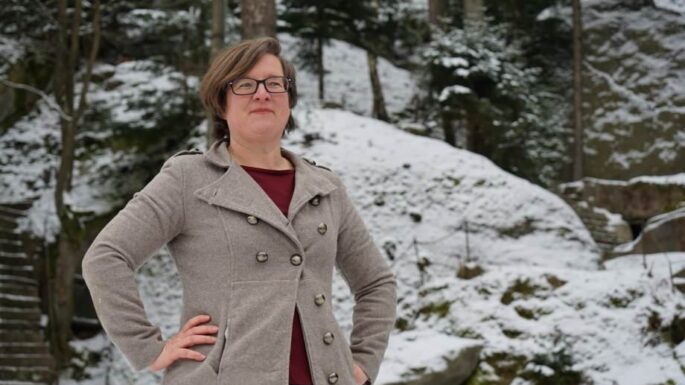 Birgit Simmler steht mit beiden Händen in den Hüften vor einer schneebedeckten hügeligen Landschaft und lächelt