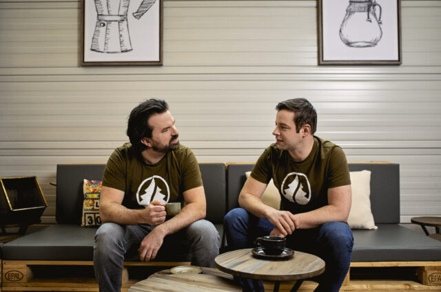 Zwei Männer sitzen auf einer Couch und schauen sich an während sie sich unterhalten und Kaffeetrinken