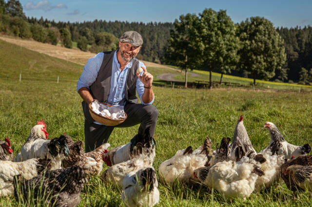 Werner kniet auf einer Wiese umgeben von vielen Hühnern und hält einen Korb mit Eiern in der Hand und ein einzelnes Ei in der anderen Hand
