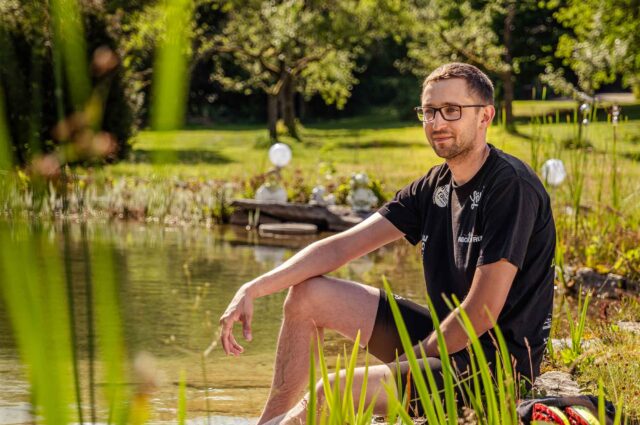 Mann mit Brille sitzt am Rande eines Sees mit kurzer Hose und Tshirt und schaut in die Ferne